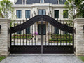 Black wrought iron gate in front of a large house with stone walls and trees.