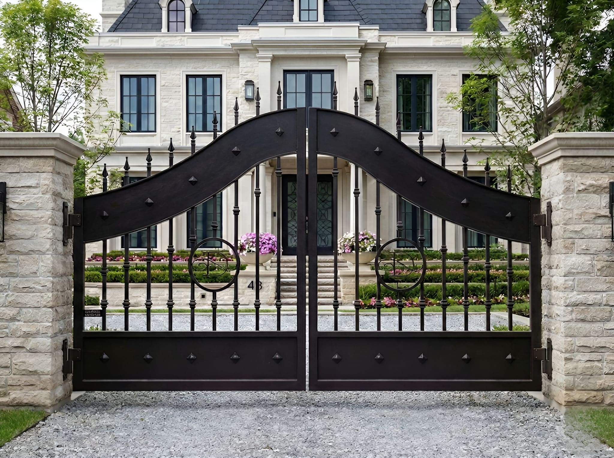 Black wrought iron gate in front of a large house with stone walls and trees.